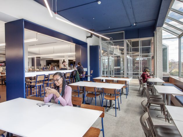 The faceted ceiling creates a raised ceiling height and allows more natural light to enter the space. The lighter, brighter tabletops further reflect the natural light. Deep blue chair legs and upholstery accents throughout the space further enforce the school’s brand.